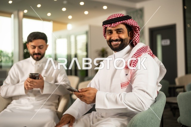 قضاء وقت ممتع في الخارج مع الأصدقاء، الترفيه والتسلية في العطلة، رجلان عربيان خليجان سعوديان يرتديان الثوب التقليدي يجلسان في المقهى بايماءات السعادة والسرور، استخدام جهاز الهاتف المحمول