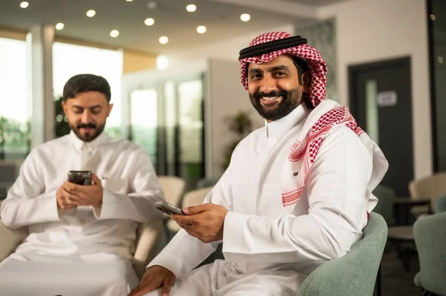 قضاء وقت ممتع في الخارج مع الأصدقاء، الترفيه والتسلية في العطلة، رجلان عربيان خليجان سعوديان يرتديان الثوب التقليدي يجلسان في المقهى بايماءات السعادة والسرور، استخدام جهاز الهاتف المحمول