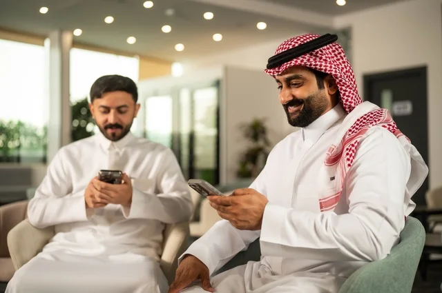 قضاء وقت ممتع في الخارج مع الأصدقاء، الترفيه والتسلية في العطلة، رجلان عربيان خليجان سعوديان يرتديان الثوب التقليدي يجلسان في المقهى بايماءات السعادة والسرور، استخدام جهاز الهاتف المحمول