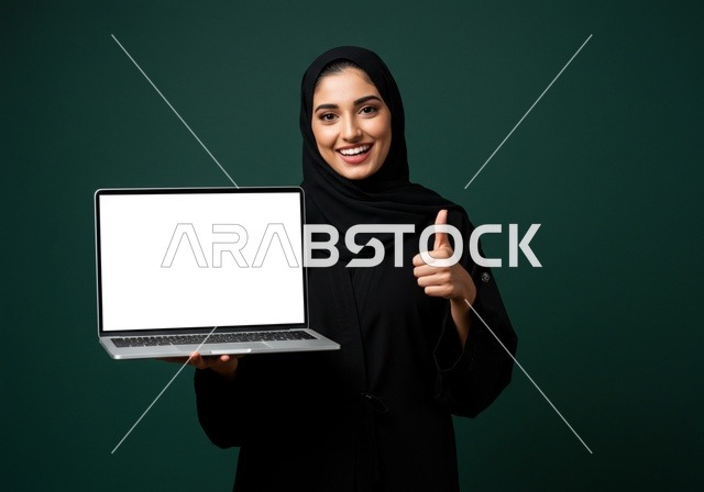 اشارة بإصبع الأبهام إلى الأعلى الأعجاب، خلفية شاشة بيضاء على حاسب محمول, النظر الى الكاميرا بتعابير السرور,  العروض والإعلانات التسويقية, بورتريه مقرب لإمرأة عربية خليجية سعودية محجبة ترتدي العباءة تمسك بيدها موبايل حديث, خلفية خضراء داكنة