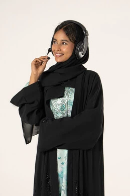 النظر الى شيء ما، الاستمتاع بسماع الأغاني، التسلية والترفيه بإستخدام تقنية الهيدفون، ارتداء سماعات الرأس الصوتية، بورترية لشابة عربية خليجية اماراتية ترتدي العباءة و الحجاب و سماعات الرأس تقوم بالاستماع للأغاني بايماءات السرور والسعاة، خلفية بيضاء