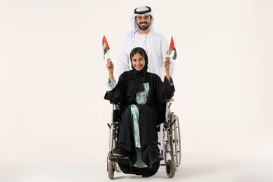 Love of the homeland and pride in national identity, national holidays and occasions, a portrait of an Arab Gulf Omani girl with special needs wearing an abaya and hijab sitting in a wheelchair alongside her husband holding the Omani flag with expressions of pride, white background.