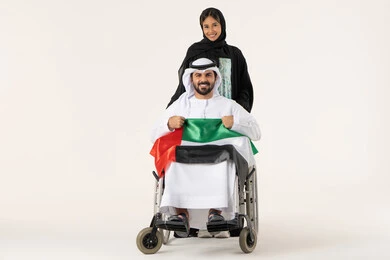 Highlighting feelings of nationalism and pride in the country, national holidays and occasions, a portrait of an Arab Gulf Omani man with special needs wearing traditional attire sitting in a wheelchair holding the Omani flag with expressions of pride alongside his wife, gestures of happiness and joy, white background.