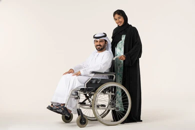 النظر الى الكاميرا بتعابير السعادة و السرور، امرأة عربية خليجية اماراتية ترتدي العباءة والحجاب تساعد زوجها على التنقل والحركة، تأمين أدوات ومعدات لمساعدة المرضي، الاخلاص والعطف بين الزوجين، جودة حياة ذوي الهمم في الامارات ، بورترية لرجل عربي خليجي اماراتي يرتدي الكندورة و الغتره مقعد يجلس على كرسي متحرك، خلفية بيضاء