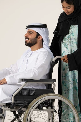 امرأة عربية خليجية اماراتية ترتدي العباءة والحجاب تساعد زوجها على التنقل والحركة، تأمين أدوات ومعدات لمساعدة المرضي، الاخلاص والعطف بين الزوجين، جودة حياة ذوي الهمم في الامارات ، بورترية لرجل عربي خليجي اماراتي يرتدي الكندورة و الغتره مقعد يجلس على كرسي متحرك، خلفية بيضاء