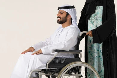الاخلاص والعطف بين الزوجين، جودة حياة ذوي الهمم في الامارات، تأمين أدوات ومعدات لمساعدة المرضي ، بورترية لرجل عربي خليجي اماراتي يرتدي الكندورة و الغتره مقعد يجلس على كرسي متحرك، امرأة عربية خليجية اماراتية ترتدي العباءة والحجاب تساعد زوجها على التنقل والحركة، خلفية بيضاء