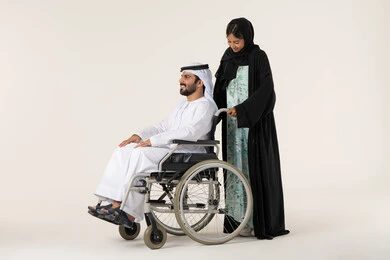 جودة حياة ذوي الهمم في الامارات، الاخلاص والعطف بين الزوجين، تأمين أدوات ومعدات لمساعدة المرضي ، بورترية لرجل عربي خليجي اماراتي يرتدي الكندورة و الغتره مقعد يجلس على كرسي متحرك، امرأة عربية خليجية اماراتية ترتدي العباءة والحجاب تساعد زوجها على التنقل والحركة، خلفية بيضاء