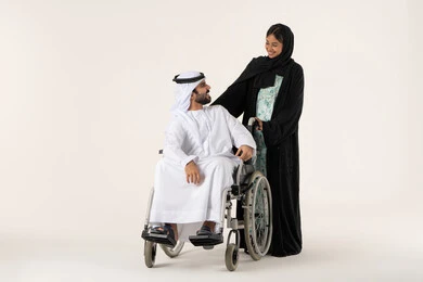 The loyalty and mutual affection between spouses, caring for people with determination and securing their needs, the lifestyle of individuals with mobility disabilities, providing tools and equipment to assist patients, a Gulf man with special needs sitting in a wheelchair looking at his wife, an Emirati Arab woman helping her husband to move and navigate, white background.