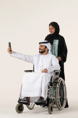 تعابير السعادة و السرور، الاهتمام بالجانب النفسي والجسدي للمقعدين، رعاية المرضى والمعاقين، تأمين معدات تساعد بالحركة والتنقل، التقاط صور سيلفي تذكارية باستخدام الجوال، بورتريه لزوجة عربية خليجية اماراتية ترتدي العباءة و الحجاب  تقوم بمساعدة زوجها من ذوي الاحتياجات الخاصه، خلفية بيضاء