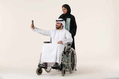 الاهتمام بالجانب النفسي والجسدي للمقعدين، رعاية المرضى والمعاقين، تأمين معدات تساعد بالحركة والتنقل، التقاط صور سيلفي تذكارية باستخدام الجوال، بورتريه لزوجة عربية خليجية اماراتية ترتدي العباءة و الحجاب  تقوم بمساعدة زوجها من ذوي الاحتياجات الخاصه، خلفية بيضاء