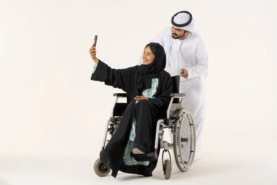 رعاية المرضى والمعاقين، تأمين معدات تساعد بالحركة والتنقل، التقاط صور سيلفي تذكارية باستخدام الجوال، بورتريه لزوج عربي خليجي اماراتي يرتدي الكندورة و الغتره يقوم بمساعدة زوجته من ذوي الاحتياجات الخاصه، الاهتمام بالجانب النفسي والجسدي للمقعدين، خلفية بيضاء