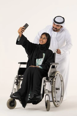 Providing equipment that assists with movement and mobility, taking commemorative selfie photos using a mobile phone, a portrait of an Emirati Gulf Arab husband wearing a kandura and ghutrah helping his wife with special needs, caring for patients and the disabled, focusing on the psychological and physical aspects of the disabled, white background.