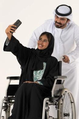 التقاط صور سيلفي تذكارية باستخدام الجوال، تأمين معدات تساعد بالحركة والتنقل، بورتريه لزوج عربي خليجي اماراتي يرتدي الكندورة و الغتره يقوم بمساعدة زوجته من ذوي الاحتياجات الخاصه، رعاية المرضى والمعاقين، الاهتمام بالجانب النفسي والجسدي للمقعدين، خلفية بيضاء