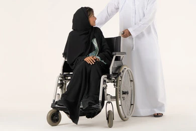 نمط حياة ذوي الإعاقة الحركية، العنايه بأصحاب الهمم وتأمين احتياجاتهم، تأمين أدوات ومعدات لمساعدة المرضي ، امراة خليجيه من ذوي الاحتياجات الخاصه تجلس على كرسي متحرك تنظر الى زوجها، رجل عربي خليجي اماراتي يرتدي الكندورة و الغتره  يساعد زوجته على التنقل والحركة ، خلفية بيضاء