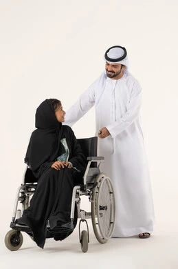 العنايه بأصحاب الهمم وتأمين احتياجاتهم ، نمط حياة ذوي الإعاقة الحركية ، تأمين أدوات ومعدات لمساعدة المرضي ، امراة خليجيه من ذوي الاحتياجات الخاصه تجلس على كرسي متحرك تنظر الى زوجها، رجل عربي خليجي اماراتي يرتدي الكندورة و الغتره يساعد زوجته على التنقل والحركة ، خلفية بيضاء
