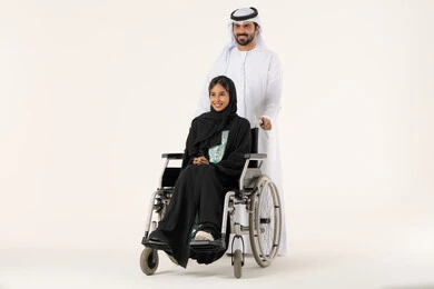 Helping patients with mobility disabilities to move easily, looking at something with expressions of happiness and joy, improving the mental state of people of determination, loyalty and compassion between spouses, a portrait of an Emirati Gulf Arab woman with special needs wearing an abaya and hijab sitting in a wheelchair accompanied by her husband.