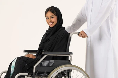 التحسين من نفسية أصحاب الهمم، الإخلاص والعطف بين الأزواج , النظر الى الكاميرا بتعابير السعادة و السرور ، مساعدة مرضى ذوي الإعاقة الحركية على التنقل بسهولة, بورترية من الجانب لامراة عربيه خليجيه اماراتية من ذوى الاحتياجات الخاصة ترتدي العباءة و الحجاب تجلس على كرسي متحرك برفقه زوجها