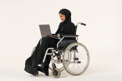 Remote business management, completing tasks via laptop, using modern devices and technologies, economics and financial matters, a side portrait of an Arab Gulf Emirati woman with special needs wearing an abaya and hijab sitting in a wheelchair working on a laptop, white background.
