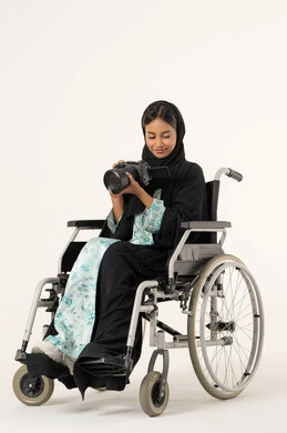 احتراف فن التصوير الفوتوغرافي، ممارسة هواية التصوير بتعابير السرور، استخدام الأجهزة التقنية الحديثة والتكنولوجيا، استخدام كاميرات رقمية عالية الدقة، بورترية لامراة عربيه خليجيه اماراتية من ذوى الاحتياجات الخاصة ترتدي العباءة و الحجاب تجلس على كرسي متحرك تقوم بالتقاط صورة تذكارية عبر كاميرا فوتوغرافية ، خلفية بيضاء