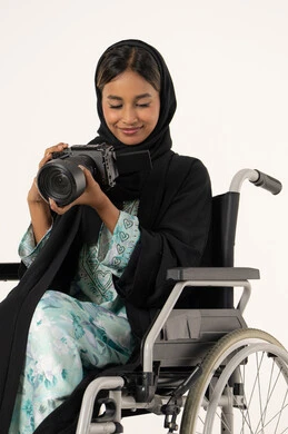 Practicing the hobby of photography with expressions of joy, using modern technical devices and technology, using high-resolution digital cameras, a portrait of an Emirati Arab woman with special needs wearing an abaya and hijab sitting in a wheelchair taking a commemorative photo with a camera, mastering the art of photography, white background.