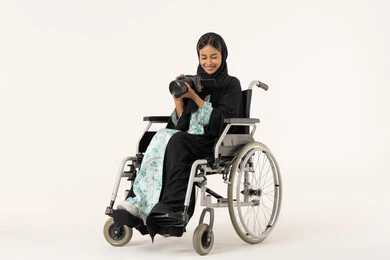 The use of modern technical devices and technology, the use of high-resolution digital cameras, a portrait of an Arab Gulf Emirati woman with special needs wearing an abaya and hijab sitting in a wheelchair taking a commemorative photo with a camera, practicing the hobby of photography with expressions of joy, mastering the art of photography, white background.