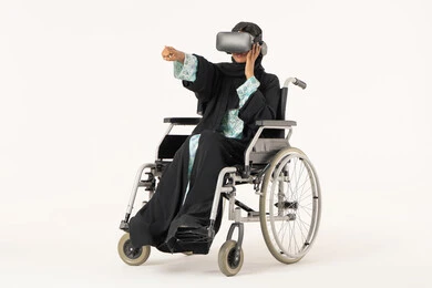 الاشارة الى شيء ما، المتعة والتسلية بالمشاهدة عبر نظارة الواقع الافتراضي، بورترية لشابة عربيه خليجيه اماراتية من ذوى الاحتياجات الخاصة ترتدي العباءة و الحجاب ونظارات ثلاثية الأبعاد (الفي ار) تجلس على كرسي متحرك, الاندماج ومحاكاة العالم الخيالي، التطور التقني والتكنولوجي ، خلفية بيضاء