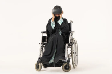 Integration and simulation of the imaginary world, the enjoyment of entertainment viewing with virtual reality technologies, a portrait of a young Arab Gulf Emirati woman with special needs wearing an abaya and hijab sitting in a wheelchair and wearing 3D glasses (VR), technological and technical development, white background.