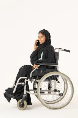 Enjoying exchanging conversations and news, making a voice phone call via mobile, a side portrait of an Emirati Arab woman with special needs wearing an abaya and hijab sitting in a wheelchair talking on a mobile phone, gestures of happiness in communicating with friends and relatives, white background.