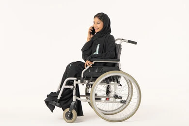 Making a voice phone call via mobile, enjoying exchanging conversations and news, a side portrait of an Arab Gulf Emirati woman with special needs wearing an abaya and hijab sitting in a wheelchair talking on a mobile phone, gestures of happiness in communicating with friends and relatives, white background.