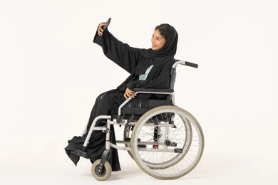 Documenting happy moments and taking commemorative photos, a side portrait of an Arab Gulf Emirati woman with special needs wearing an abaya and hijab sitting in a wheelchair holding a mobile phone taking a selfie, gestures of self-confidence, the concept of elegance and attention to appearance, white background.