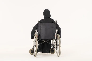 The use of wheelchairs to assist with movement and mobility, improving the quality of life for individuals with mobility disabilities, a portrait from behind of an Arab Gulf Emirati woman with special needs wearing an abaya and hijab sitting in a wheelchair, white background.