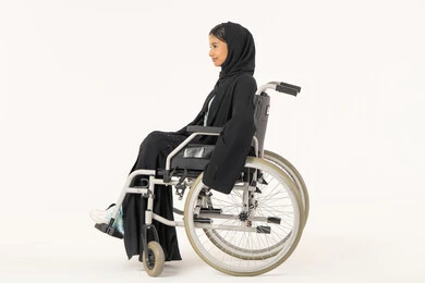 العنايه بأصحاب الهمم وتأمين احتياجاتهم الخاصة، توفير اجهزة ومعدات تساعد بالحركة والتنقل، بورتريه من الجانب لامرأة عربية اماراتية خليجية محجبة من ذوي الاحتياجات الخاصة ترتدي العباءة السوداء و تجلس على كرسي متحرك, خلفية بيضاء