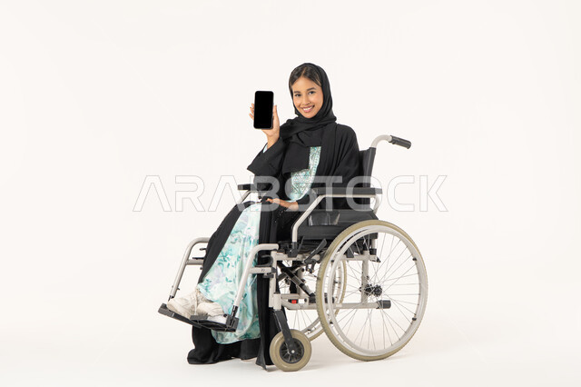 تسهيل حياة ذوي الهمم باستخدام التقنيه والتكنولوجيا, شاشة سوداء فارغه على الهاتف المحمول,  بورتريه لإمرأة عربية خليجية اماراتية خليجية محجبة مبتسمة من ذوي الاحتياجات الخاصة ترتدي العباءة السوداء و تجلس على كرسي متحرك و  تقوم بعرض برامج وتطبيقات ذوي الاحتياجات الخاصه, خلفية بيضاء