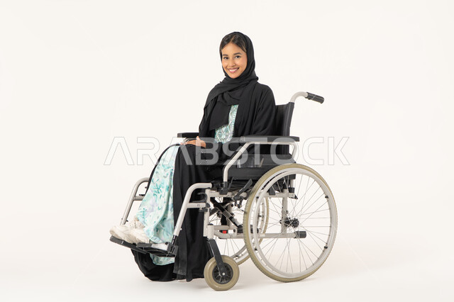 مفهوم التفاؤل وحب الحياة، العنايه بأصحاب الهمم و تأمين احتياجاتهم، توفير اجهزة ومعدات تساعد بالحركة والتنقل، بورتريه لإمرأة عربية خليجية  اماراتية خليجية محجبة من ذوي الاحتياجات الخاصة ترتدي العباءة السوداء و تجلس على كرسي متحرك و تنظر للكاميرا بابتسامة, خلفية بيضاء