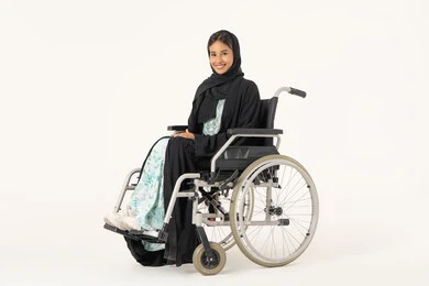 مفهوم التفاؤل وحب الحياة، العنايه بأصحاب الهمم و تأمين احتياجاتهم، توفير اجهزة ومعدات تساعد بالحركة والتنقل، بورتريه لإمرأة عربية خليجية  اماراتية خليجية محجبة من ذوي الاحتياجات الخاصة ترتدي العباءة السوداء و تجلس على كرسي متحرك و تنظر للكاميرا بابتسامة, خلفية بيضاء