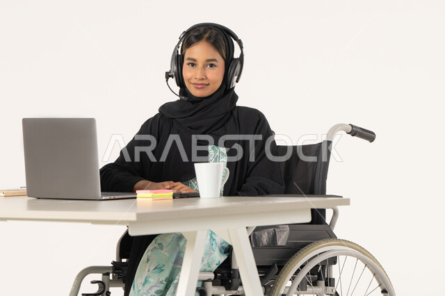 ادارة الأعمال عن بعد، بورتريه لامرأة عربية اماراتية خليجية محجبة مبتسمة تنظر الى الكاميرا من ذوي الاحتياجات الخاصة ترتدي العباءة السوداء و تجلس على كرسي متحرك أمام جهاز اللابتوب و تضع سماعات الهيدفون, مهن ووظائف ادارية مكتبية, خلفية بيضاء