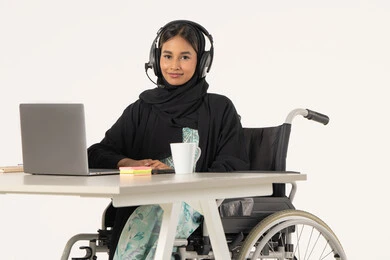 Remote business management, portrait of a veiled, smiling, Arab Emirati Gulf woman looking at the camera, with special needs, wearing a black abaya, sitting on a wheelchair in front of a laptop and wearing headphones, office administrative professions and jobs, white background