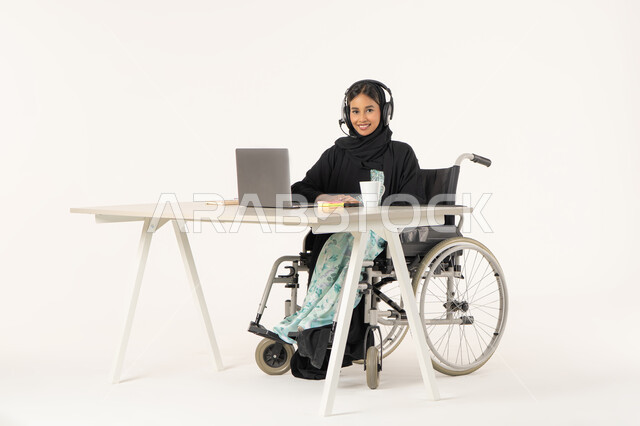 العمل من خلال النترنت, ادارة الأعمال عن بعد، بورتريه لامرأة عربية اماراتية خليجية محجبة مبتسمة تنظر الى الكاميرا من ذوي الاحتياجات الخاصة ترتدي العباءة السوداء و تجلس على كرسي متحرك أمام جهاز اللابتوب و تضع سماعات الهيدفون, مهن ووظائف ادارية مكتبية, خلفية بيضاء