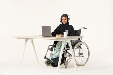 العمل من خلال النترنت, ادارة الأعمال عن بعد، بورتريه لامرأة عربية اماراتية خليجية محجبة مبتسمة تنظر الى الكاميرا من ذوي الاحتياجات الخاصة ترتدي العباءة السوداء و تجلس على كرسي متحرك أمام جهاز اللابتوب و تضع سماعات الهيدفون, مهن ووظائف ادارية مكتبية, خلفية بيضاء