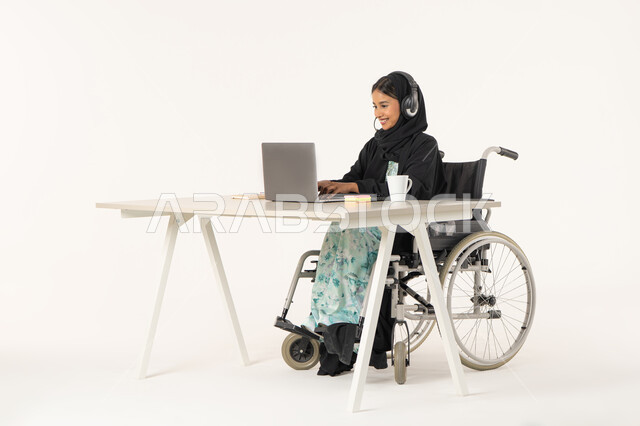 النقر على لوحة المفاتيح, جودة حياة ذوي الهمم في الامارات, بورتريه لامرأة عربية اماراتية خليجية محجبة مبتسمة من ذوي الاحتياجات الخاصة ترتدي العباءة السوداء و تجلس على كرسي متحرك و أمامها جهاز اللابتوب, خلفية بيضاء