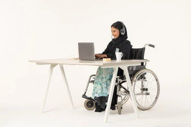 Clicking on the keyboard, quality of life for people with disabilities in the Emirates, portrait of a smiling veiled Emirati Gulf Arab woman with special needs wearing a black abaya and sitting on a wheelchair with a laptop in front of her, white background