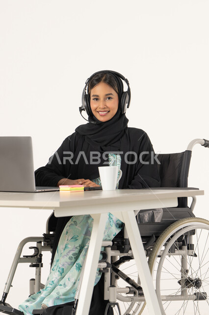 ابتسامة ونظرة إلى الكاميرا، تأمين أدوات ومعدات لمساعدة المرضي، جودة حياة ذوي الهمم في الامارات,  بورتريه لامرأة عربية اماراتية خليجية محجبة مبتسمة تنظر الى الكاميرا من ذوي الاحتياجات الخاصة ترتدي العباءة السوداء و تجلس على كرسي متحرك أمام جهاز اللابتوب و تضع سماعات الهيدفون, مهن ووظائف ادارية مكتبية, خلفية بيضاء