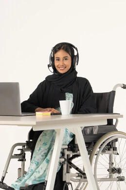 Smile and look at the camera, providing tools and equipment to help patients, quality of life for people with disabilities in the Emirates, portrait of a smiling veiled Emirati Gulf Arab woman looking at the camera with special needs, wearing a black abaya and sitting on a wheelchair in front of a laptop and wearing headphones, office administrative professions and jobs, white background