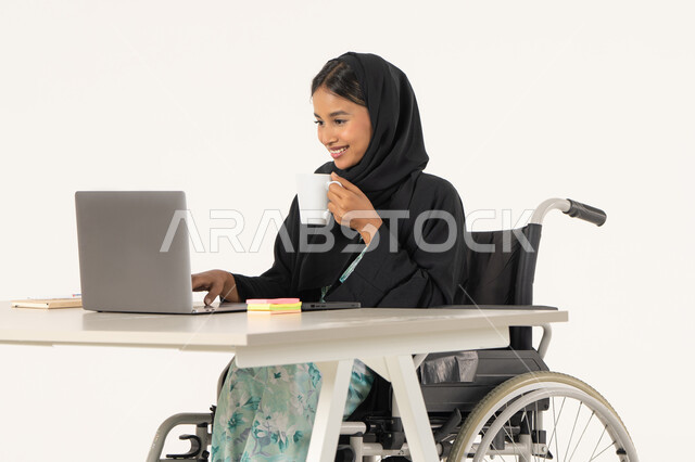 بيئة عمل مريحة في الشركات الاماراتية, بورتريه لامرأة عربية اماراتية خليجية محجبة من ذوي الاحتياجات الخاصة ترتدي العباءة السوداء و تجلس على كرسي متحرك و تباشر عملها من خلال اللابتوب و تشرب مشروب دافئ, خلفية بيضاء