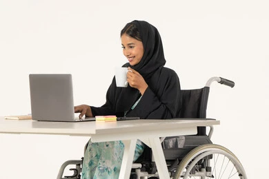 بيئة عمل مريحة في الشركات الاماراتية, بورتريه لامرأة عربية اماراتية خليجية محجبة من ذوي الاحتياجات الخاصة ترتدي العباءة السوداء و تجلس على كرسي متحرك و تباشر عملها من خلال اللابتوب و تشرب مشروب دافئ, خلفية بيضاء