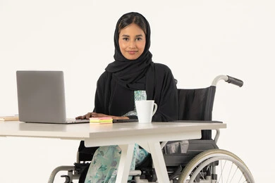 Integrating technology and advanced technology into work, office profession and job, portrait of a veiled Emirati Gulf Arab woman smiling looking at the camera with special needs wearing a black abaya and sitting on a wheelchair in front of a laptop, white background