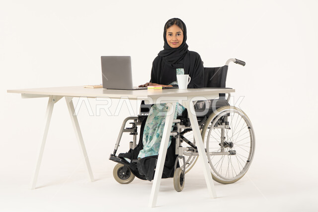 Using a laptop for business, integrating technology and advanced technology into work, office profession and job, portrait of a veiled, smiling, Arab, Emirati, Gulf woman with special needs looking at the camera, wearing a black abaya and sitting in a wheelchair in front of a laptop, white background