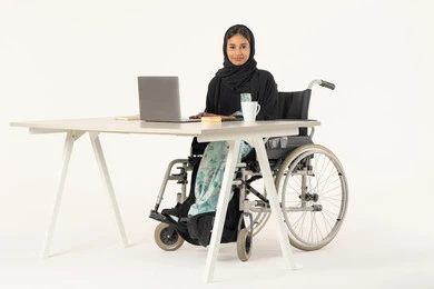 استعمال اللابتوب لادارة الأعمال, دمج التقنية والتكنولوجيا المتطورة بالعمل، مهنة ووظيفة مكتبية، بورتريه لامرأة عربية اماراتية خليجية محجبة مبتسمة تنظر الى الكاميرا من ذوي الاحتياجات الخاصة ترتدي العباءة السوداء و تجلس على كرسي متحرك أمام جهاز الحاسب المحمول, خلفية بيضاء