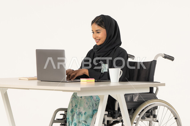 استخدام اللابتوب لانجاز المهام والعمل، جودة حياة ذوي الهمم في الامارات, بورتريه لامرأة عربية اماراتية خليجية محجبة من ذوي الاحتياجات الخاصة ترتدي العباءة السوداء و تجلس على كرسي متحرك و تقوم بالنقر على لوحة المفاتيح عبر جهاز الحاسوب المحمول, خلفية بيضاء