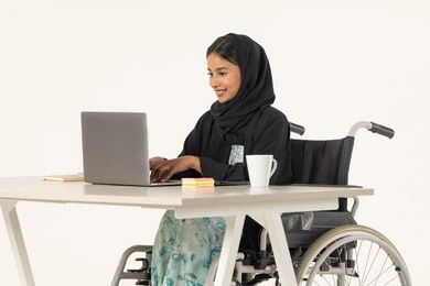 استخدام اللابتوب لانجاز المهام والعمل، جودة حياة ذوي الهمم في الامارات, بورتريه لامرأة عربية اماراتية خليجية محجبة من ذوي الاحتياجات الخاصة ترتدي العباءة السوداء و تجلس على كرسي متحرك و تقوم بالنقر على لوحة المفاتيح عبر جهاز الحاسوب المحمول, خلفية بيضاء